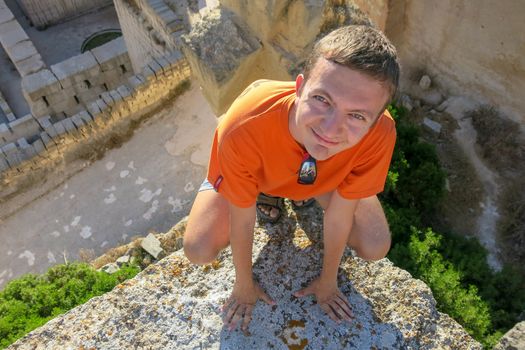 A young man in a hiking outfit squats at the edge of a steep cliff. Yang caucasian man squatting on a rock edge. Top view to boy over deep abyss. Old Stone-pit on Menorca, Balearic Islands, Spain.