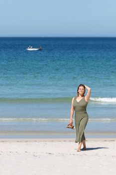 Middle-aged blonde woman in a green dress walking barefoot on the beach on the beach with her sandals in one hand