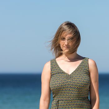 Close-up of a middle-aged blonde woman in a dress and hair shaking in the wind, with the sea in the background.