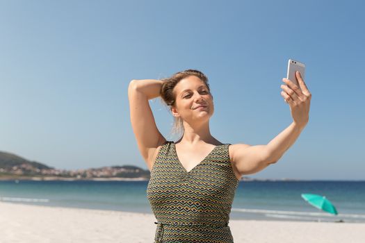 Middle-aged blonde woman in a green dress making a selfie on the beach with the sea in the background