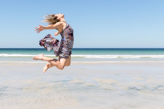 Middle-aged blonde woman in a green dress jumping with her arms open on the beach