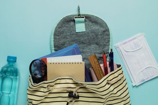 Learning supplies with hygiene face mask and fresh drinking water in a student backpack on blue background for education and back to school in an outbreak of plague situation concept