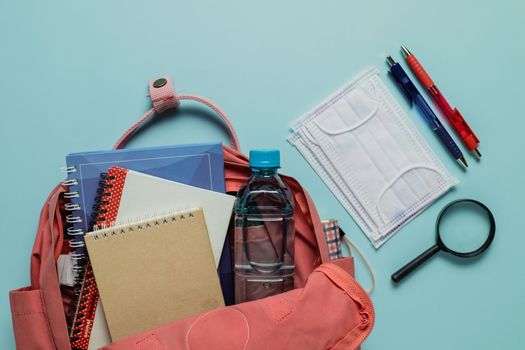 Learning supplies with hygiene face mask in a pink student backpack on blue background for education and back to school in an outbreak of plague situation concept