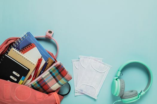 Learning supplies with hygiene face mask and headphones in a pink student backpack on blue background for education and back to school in an outbreak of plague situation concept