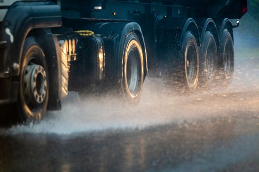 Rain water splash flow from wheels of heavy truck moving fast in daylight cit. Truck moving on asphalt road during heavy summer storm rain. Close-up with selective focus and blur.