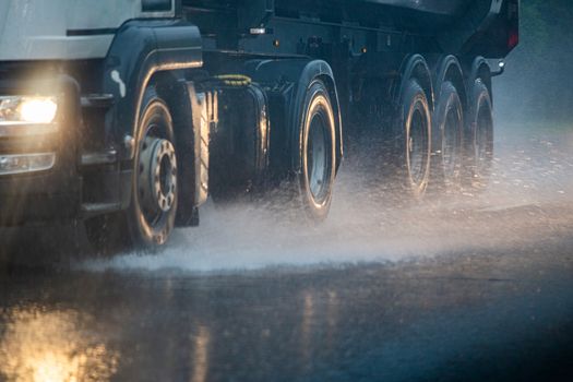 Rain water splash flow from wheels of heavy truck moving fast in daylight cit. Truck moving on asphalt road during heavy summer storm rain. Close-up with selective focus and blur.