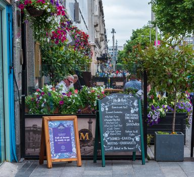 Dublin, Ireland --July 9, 2018. Patrons are seated at an outdoor Bar & Grill in Dublin, surrounded by flowers as pedestrians pass by.