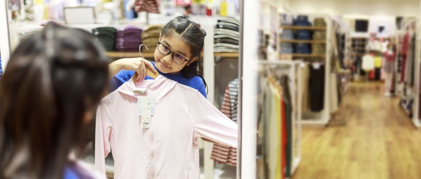 Little girl tried clothes in front of mirror in shopping mall. Asian girl smileing happy to choice and tried clothes in front of mirror in shop with copy space banner. Girl holding  clothes for tried.