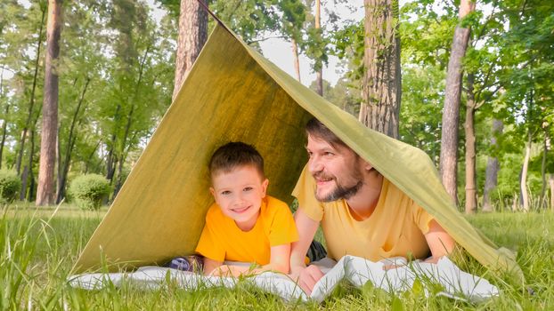 Smiling father with son in tent on green grass having fun. Family vacation in nature in the park. Father and son communicate in a house with a blanket. Travel close to home. Childhood concept.