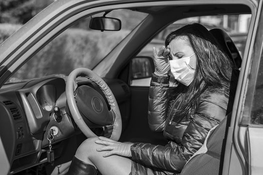 a girl in a car in a medical anti virus mask with poor health holds her head with her hand