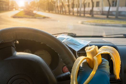 food, a banana in the hand of a driver behind the wheel of a car in rubber gloves and against the background of a medical anti-virus mask. Safe food