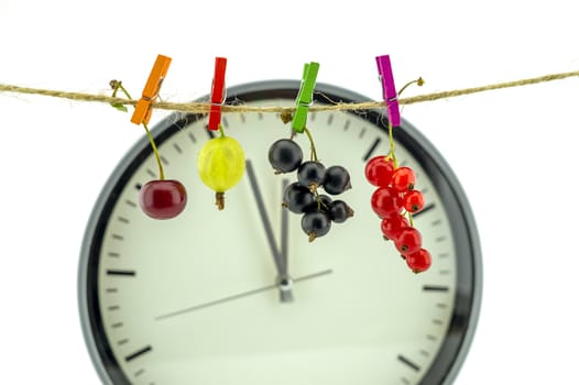 Clock showing five minutes to twelve with fresh a cherry, gooseberry, red and black currants hanging on a line from clothes pegs in a healthy diet concept with copy space over white