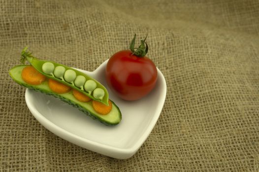 Healthy fresh vegetable sandwich with carrots, peas in a pod and cucumber balance on top of each and red ripe tomato in a heart-shaped plate on jute