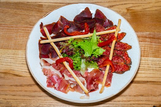 Plate with delicacies. Trio of dried delicacies. Basturma, prosciutto, sausage flat. White plate. Wooden table