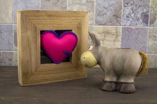 Cute little toy donkey look at handcrafted pink heart in a wooden frame against a tiled background