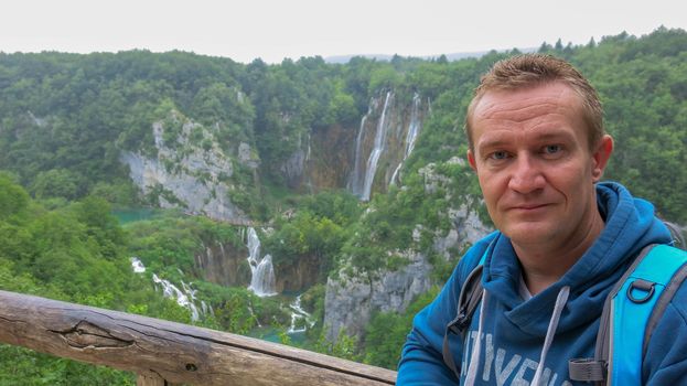 Blue-eyed tourist in a blue hoodie. A middle-aged man leans against a fence and enjoys the view in Plivice lakes National Park, Croatia.