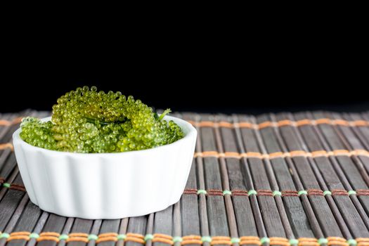 Fresh sea grapes or caviar seaweed in white bowl on wooden mat and black background. Healthy food concept.