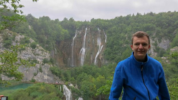 A young man leans on a wooden railing. In the background waterfalls in Plitvice Lakes National Park. Teenager in a blue hoodie stands against a waterfall.