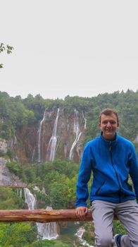 A young man leans on a wooden railing. In the background waterfalls in Plitvice Lakes National Park. Teenager in a blue hoodie stands against a waterfall.