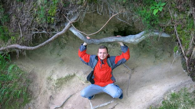 Teenager sitting on the roots of a tree. Young man in a red jacket and panties climbs between the roots of a tree. The guy is resting under a big tree. The guy is sitting on the roots.