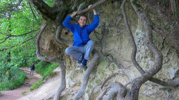 Teenager sitting on the roots of a tree. Young man in a blue hoodie and panties climbs between the roots of a tree. The guy is resting under a big tree. The guy is sitting on the roots.