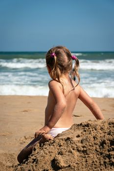 Baby girl in summertime rear view. Girl child relax alone at seaside. Preschool blonde looking at the ocean waves. Adorable little kid sitting back at pile of sand. Pensive child at seashore scenery. 