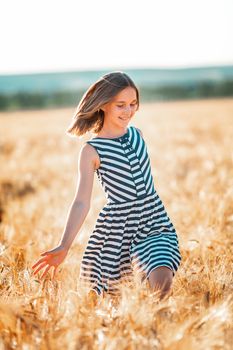 Happy teenage beautiful girl running down golden wheat field at the sunset