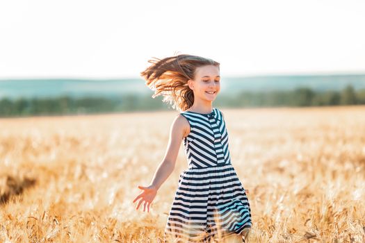 Happy teenage beautiful girl running down golden wheat field at the sunset