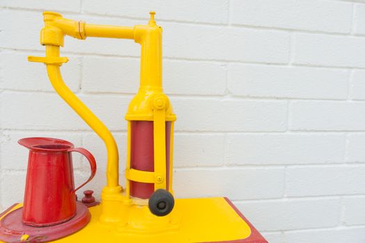 Bright red oil can on yellow old-fashioned oil hand pump closeup against white wall