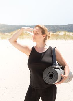 Woman in sportswear with a rolled-up mat under her arm at the beach, and protecting herself from the sun with one hand to look away