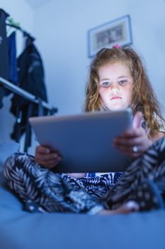 Focused small cute girls using digital tablet in bed, watching movie or play game in evening.