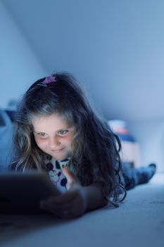 Smiling happy cute girl with long curly golden hair using digital tablet in bed, watching movie or play game in evening.