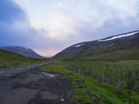 Asphalt road curve through empty northern landscape with green grass colorful hills and sunset dramatic sky, way to the mountains in Iceland western highlands, copy space