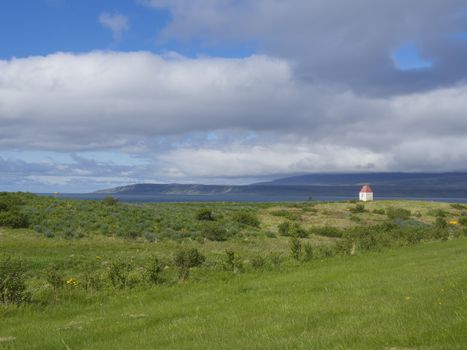 Iceland summer landscape with lush green grass meadow, sea and cliffs on horizon, small house and blue sky white clouds background