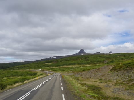 asphalt road curve in landscape with hat shape mountains with snow capes, green hills and grass meadow, blue sky white clouds, Iceland summer
