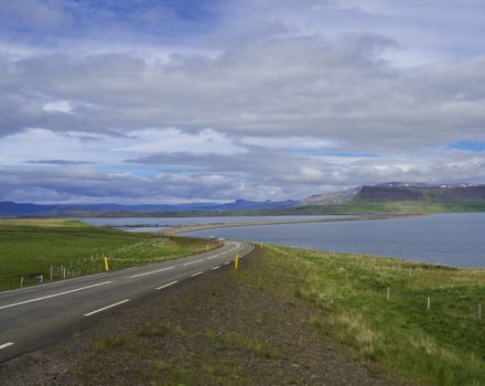 Asphalt road line through rural north summer landscape with green grass. colorful cliffs, lake, blue mountains and dramatic sky, Iceland western fjords, golden hour light.