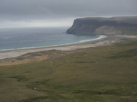 view on pink beige sand beach and green grass shore in iceland west fjords with green cliff and ocean, moody sky background