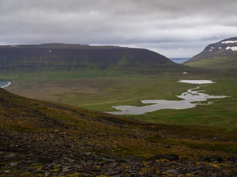 Northern summer landscape, beautiful snow covered cliffs and fljotsvatn lake in Fljotavik cove in Hornstrandir, west fjords, Iceland, with river stream, green grass meadow, moody sky background