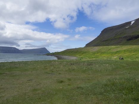 View on adalvik cove with rusty traktor and summer cottages at latrar in west fjords nature reserve Hornstrandir in Iceland, with green grass meadow, stone beach, ocean, snow patched hills and blue sky clouds background