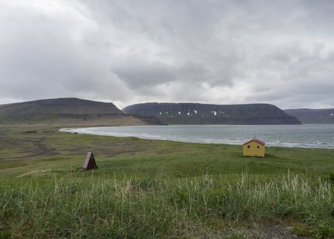 View on latrar camp site in adalvik cove with yellow emergency shelter cabin in west fjords nature reserve Hornstrandir in Iceland, with green grass meadow, stone beach, ocean, snow patched hills and dark clouds background