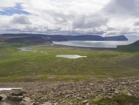 View on adalvik and latrar in west fjords nature reserve Hornstrandir in Iceland with lake and river stream, green grass meadow, beach, ocean, hills and dramatic cliffs, dark cloudy sky background