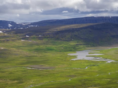 Northern summer landscape, View on beautiful cliffs in adalvik cove in west fjords nature reserve Hornstrandir in Iceland, with river stream delta, red house, snow patched green meadow and dark clouds background