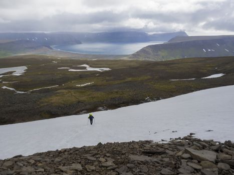 Lonely man hiker with heavy backpack walking on snow field in west fjords nature reserve Hornstrandir in Iceland with view on adalvik cove, ocean, hills and dramatic cliffs, dark cloudy sky background