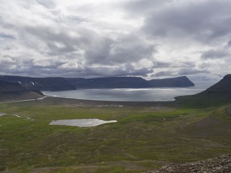 View on adalvik and latrar in west fjords nature reserve Hornstrandir in Iceland with lake and river stream, green grass meadow, beach, ocean, hills and dramatic cliffs, dark cloudy sky background