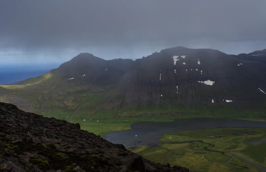Northern summer landscape, View on beautiful snow covered cliffs and fljotsvatn lake in Fljotavik cove in Hornstrandir, west fjords, Iceland, with river stream, green grass meadow, moody sky background