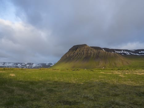 View on beautiful snow covered cliffs mountain and hills in Fljotavik cove, Hornstrandir, west fjords, Iceland, with green grass meadow, golden hour light, blue sky background.