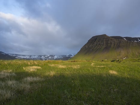 View on beautiful snow covered cliffs mountain and hills in Fljotavik cove, Hornstrandir, west fjords, Iceland, with green grass meadow, golden hour light, blue sky background