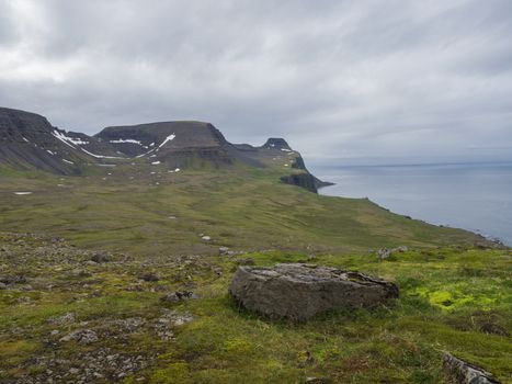 beautiful snow spotted green cliffs and blue sea horizon with big rock stone and green grass meadow, moody sky background, Hornstrandir, west fjords, Iceland