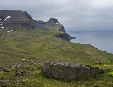 beautiful snow spotted green cliffs and blue sea horizon with big rock stone and green grass meadow, moody sky background, Hornstrandir, west fjords, Iceland