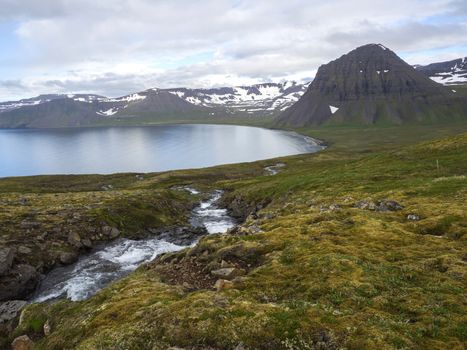 Northern summer landscape, beautiful snow covered cliffs and Alfsfell mountain, sea in Hloduvik cove with wild water stream, green meadow, clouds, in Hornstrandir, west fjords, Iceland